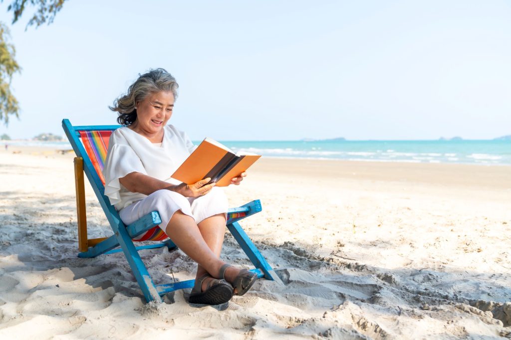 senior Asain woman sitting on a chair on the beach reading a book and relaxing through retirement