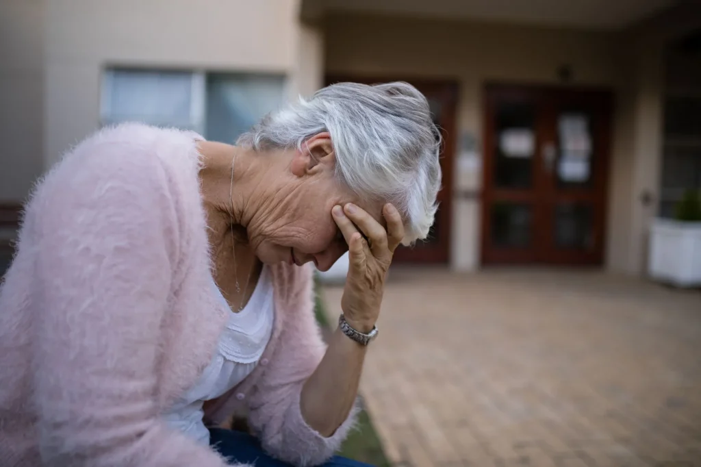 Side view of depressed senior woman sitting on bench with head in hand against nursing home or hospital feeling in disbelief