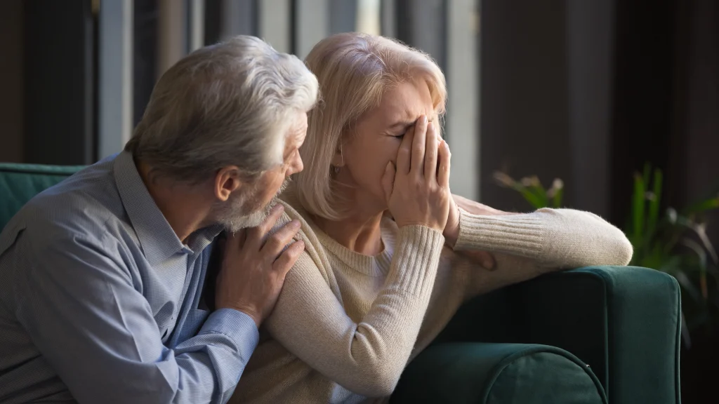 Grey haired man supporting crying unhappy mature woman, health problem, disease, bad news, loving middle aged husband hugging old wife by shoulders, sitting on couch at home,
