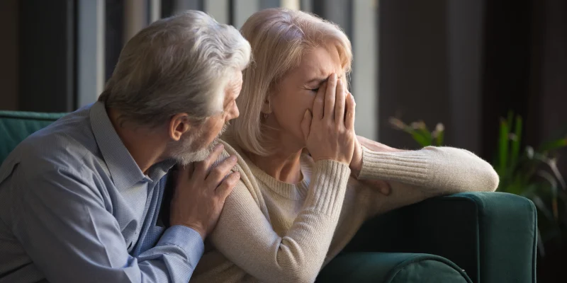 Grey haired man supporting crying unhappy mature woman, health problem, disease, bad news, loving middle aged husband hugging old wife by shoulders, sitting on couch at home,