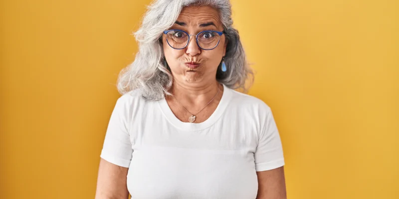 Middle age woman with grey hair standing over yellow background puffing cheeks with funny face. mouth inflated with air, crazy expression.