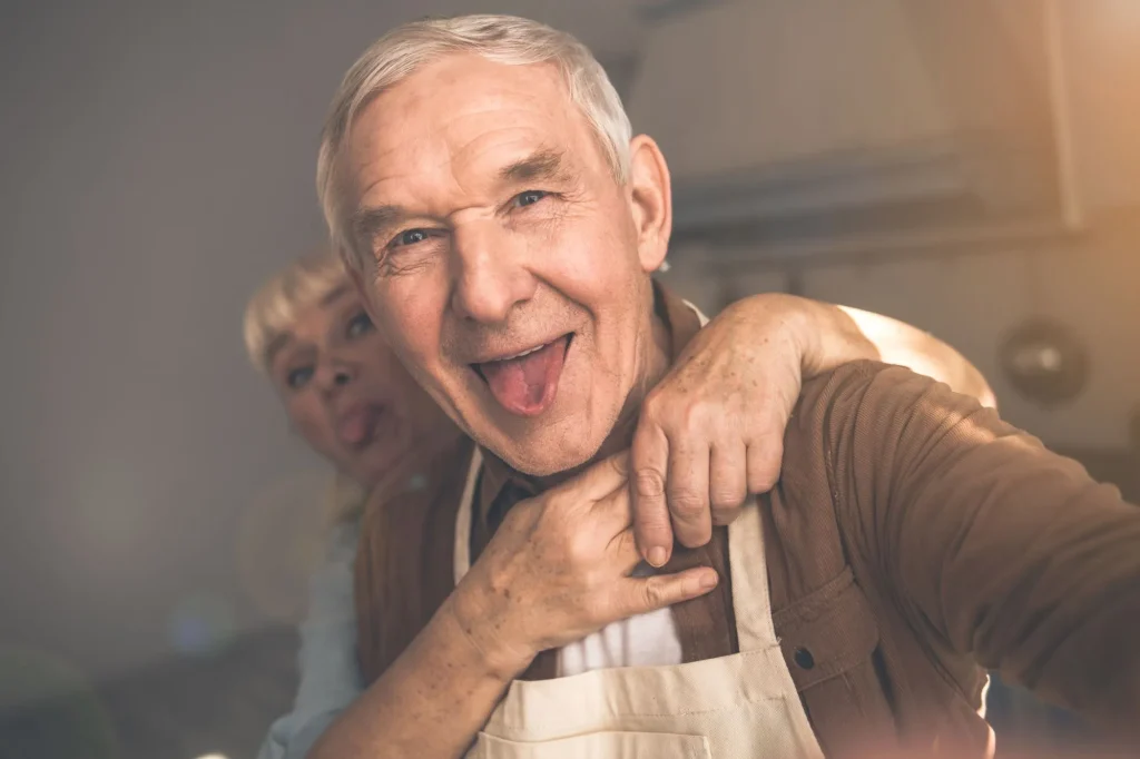 Portrait of excited mature man is making selfie with his wife. Woman is embracing husband from behind and showing tongue to camera. They are standing in kitchen
