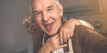 Portrait of excited mature man is making selfie with his wife. Woman is embracing husband from behind and showing tongue to camera. They are standing in kitchen