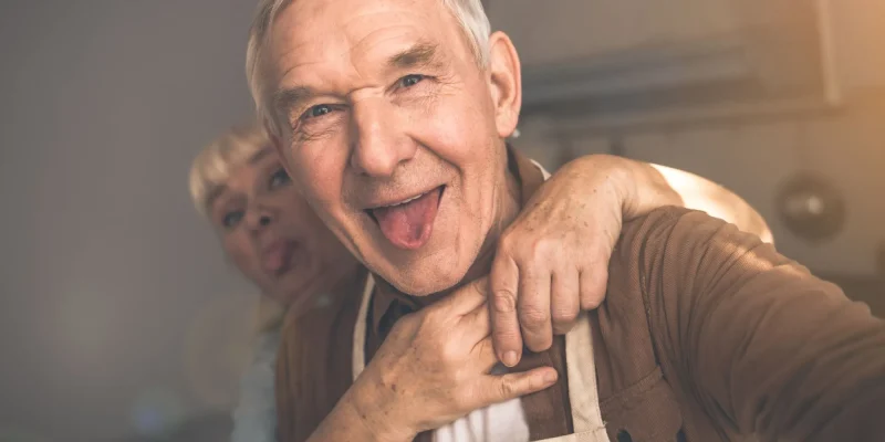 Portrait of excited mature man is making selfie with his wife. Woman is embracing husband from behind and showing tongue to camera. They are standing in kitchen