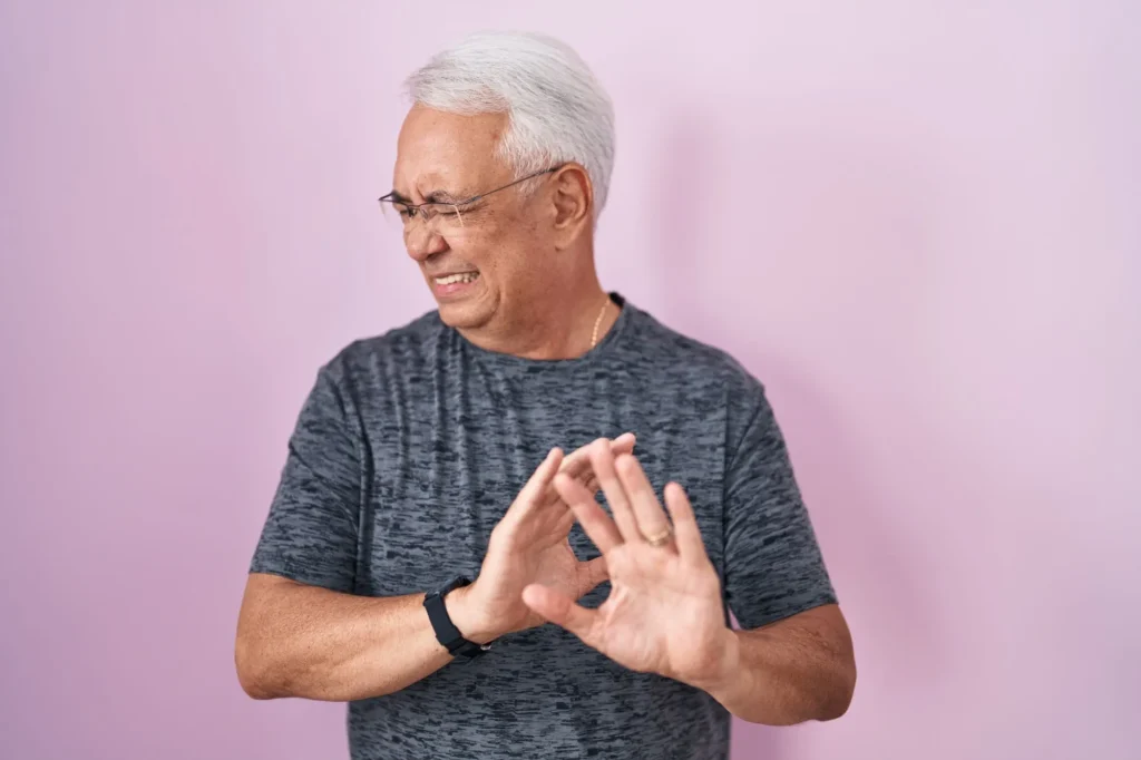 Middle age man with grey hair standing over pink background disgusted expression, displeased and fearful doing disgust face because aversion reaction, dreading his colonoscopy