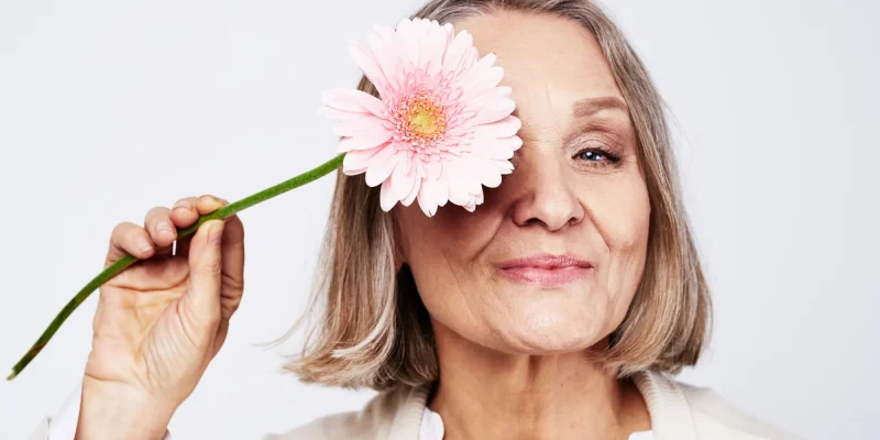 Cheerful elegant elderly woman holding a flower near the face on a gray background