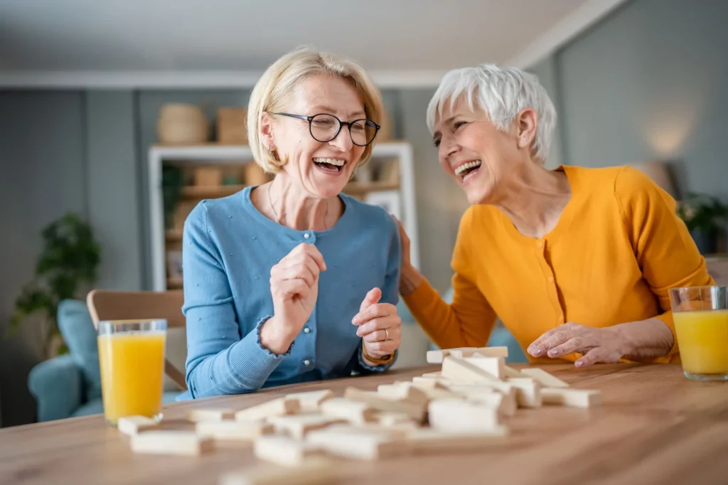 Two senior women friends sitting at a kitchen table laughing together