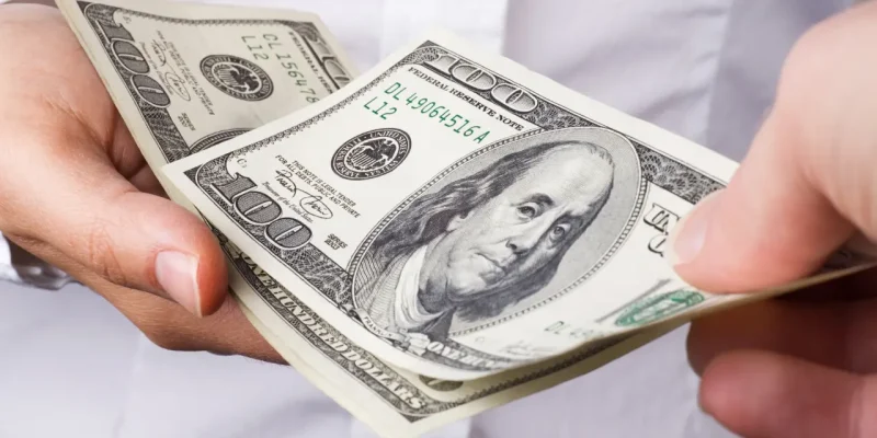 one hand handing another one-hundred dollar bills