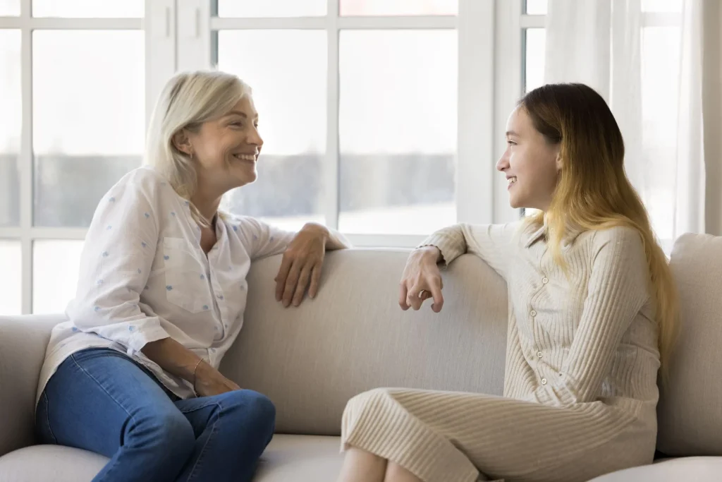 Positive teenage child and happy grandmother sitting on couch together, relaxing at home, talking, laughing, having fun. Grandma and teen granddaughter meeting at home