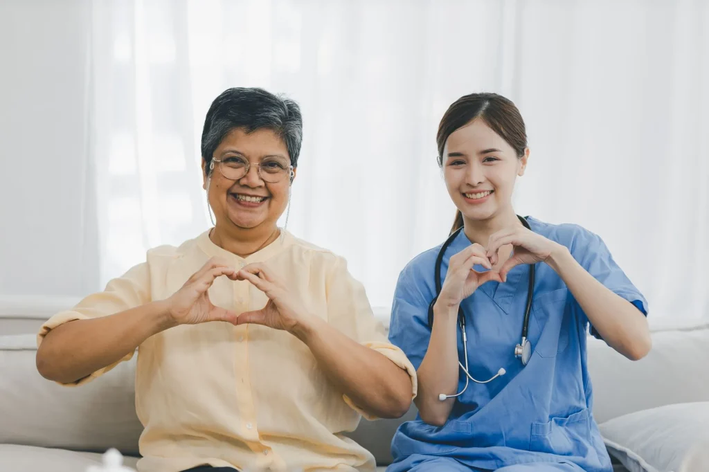 home caregiver and senior woman sitting on the couch doing heart hands