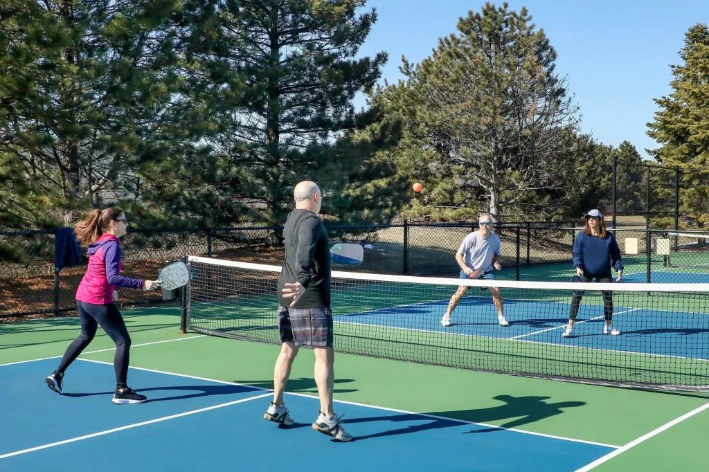 A competitive doubles game of pickleball at the net with a group of men and women on a blue and green court in spring.