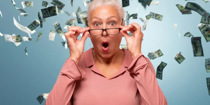 smart money senior or retiree woman with glasses down and animated look on her face on blue background with money floating
