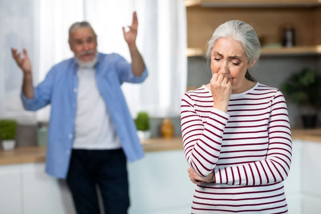 Psychological Abuse, financial abuse. Upset Senior Woman Crying In Kitchen While Arguing With Husband, Elderly Spouses Suffering Marital Crisis, Angry Man Standing On Background And Blaming Wife, Selective Focus