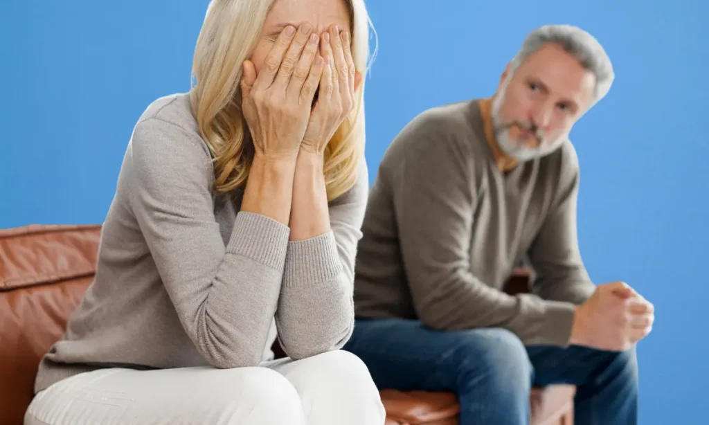 upset spouse sitting on couch with hands over face while man doesn't seem to care