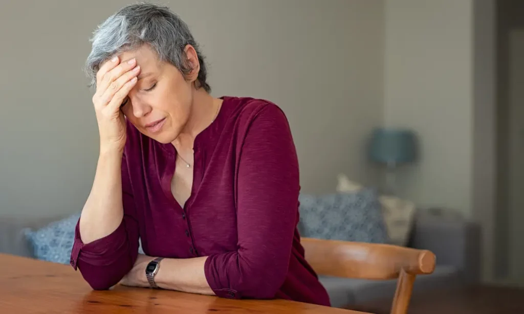 senior woman wearing magenta shirt, sitting at table with head in hand and eyes closed, feeling frustrated, bitter, upset