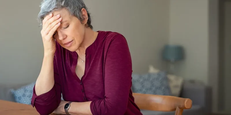 senior woman wearing magenta shirt, sitting at table with head in hand and eyes closed, feeling frustrated, bitter, upset