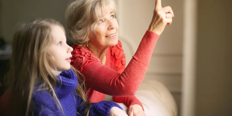 grandmother sitting by window with granddaughter pointing up and smiling