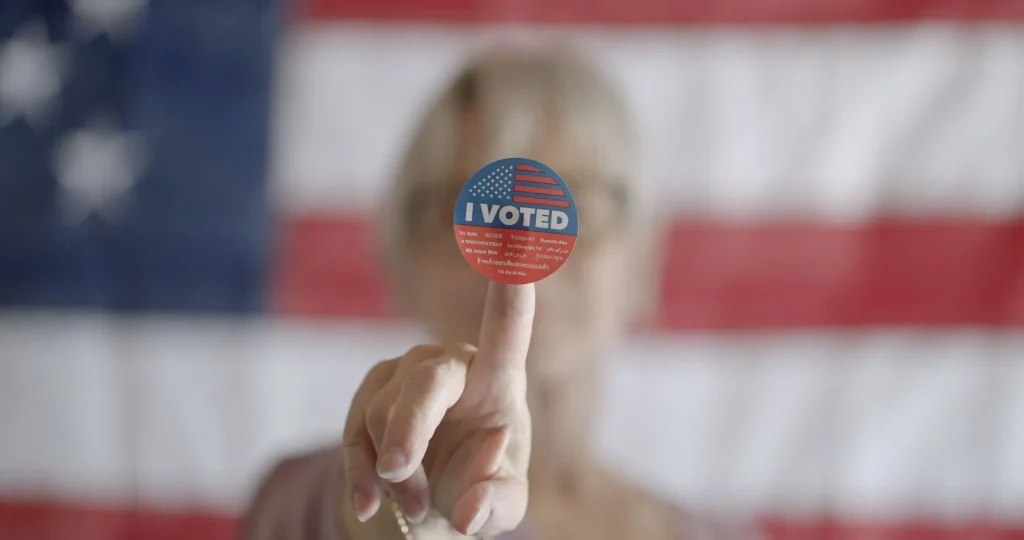 Elderly woman voter holds up sticker on finger, in front of her face. Close up, focus on sticker which says "I voted"