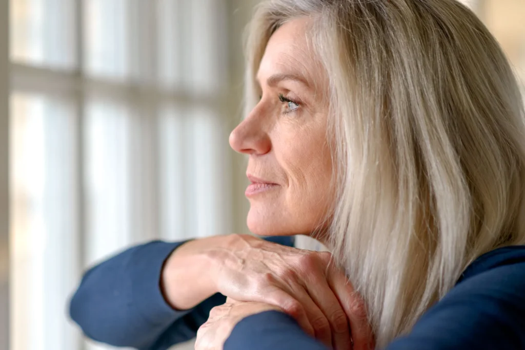 Attractive thoughtful woman with serious expression standing with her hand to her chin staring quietly out of a large window