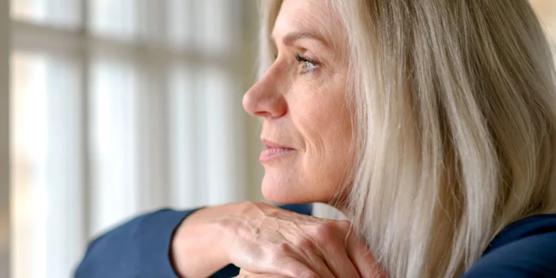 Attractive thoughtful woman with serious expression standing with her hand to her chin staring quietly out of a large window