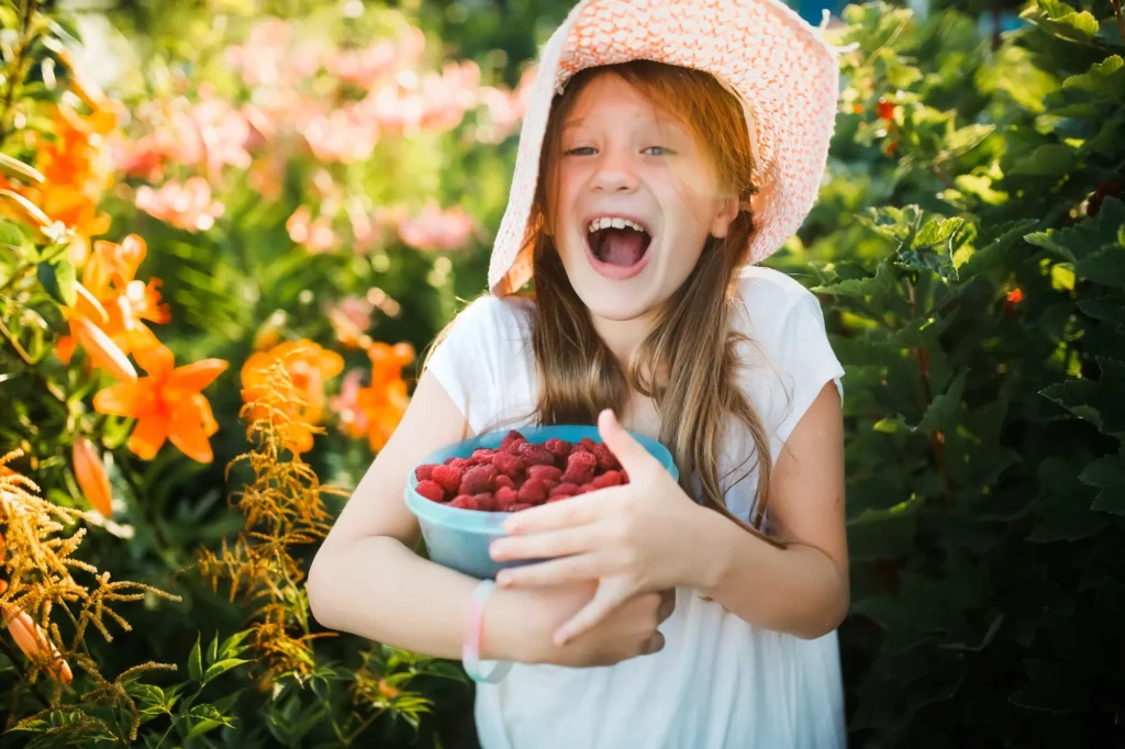 little girl, tween, picking berries outside wearing a hat and smiling big, red hair