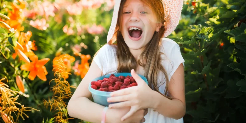 little girl, tween, picking berries outside wearing a hat and smiling big, red hair