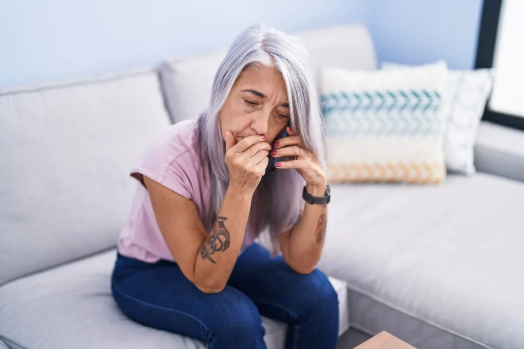 Middle age grey-haired woman talking on smartphone with worried expression at home