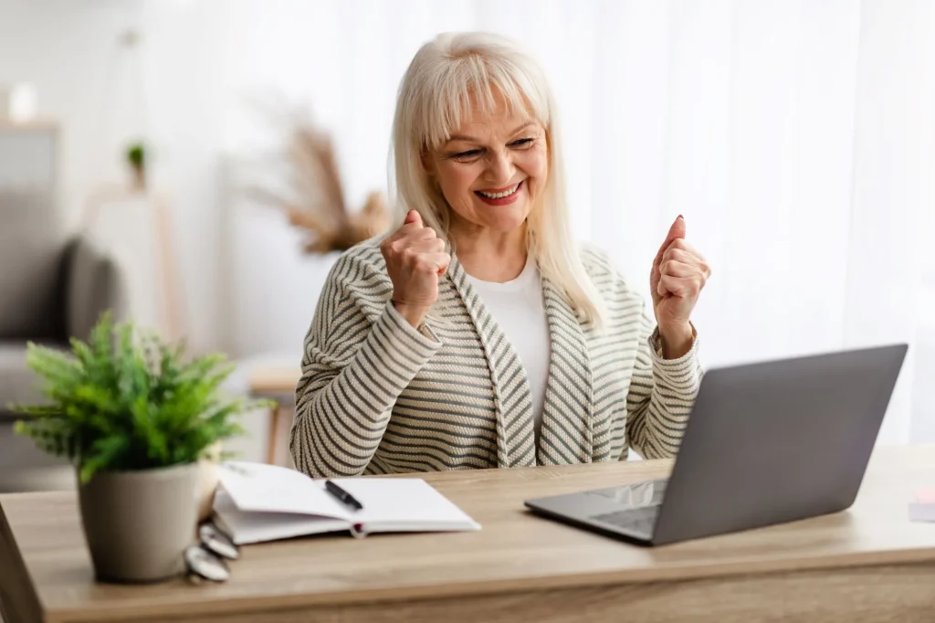 happy smiling senior woman sitting at desk celebrating with hands up at computer screen