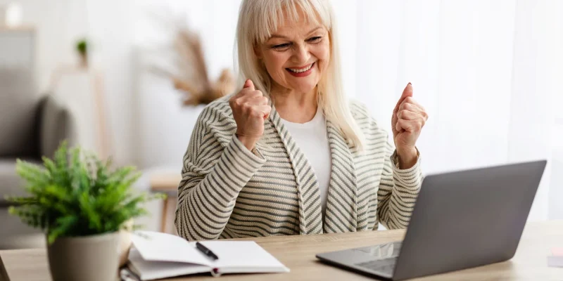 happy smiling senior woman sitting at desk celebrating with hands up at computer screen