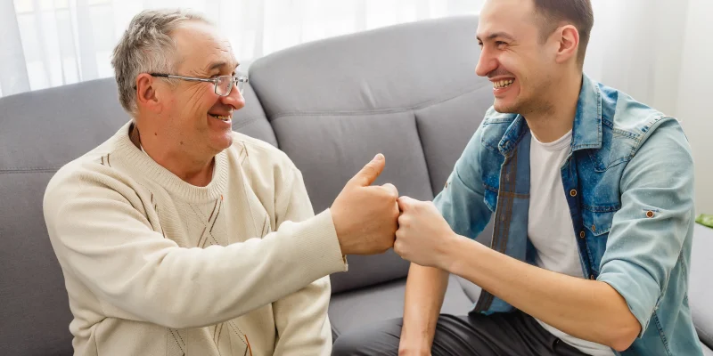 senior man and college student man sitting on living room couch smiling and doing a fist bump
