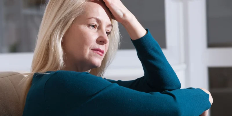 Sad depressed woman at home sitting on the couch, looking down and touching her forehead, loneliness and pain concept