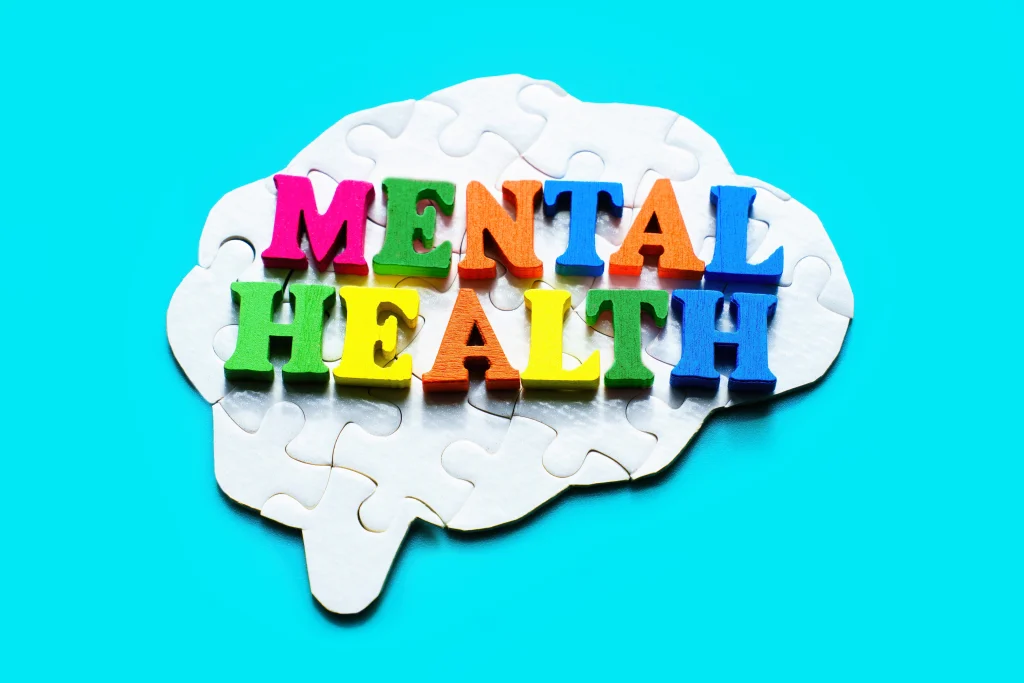 Assembled brain-shaped jigsaw puzzle on a light blue background with the words "Mental Health" spelled out in colorful wooden letters. Mental well-being and addressing mental health issues.