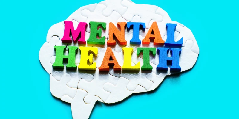 Assembled brain-shaped jigsaw puzzle on a light blue background with the words "Mental Health" spelled out in colorful wooden letters. Mental well-being and addressing mental health issues.