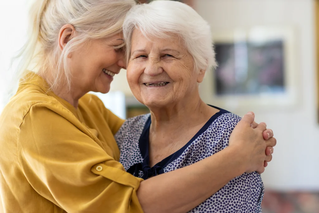 adult child daughter wearing a yellow shirt, smiling and hugging elderly senior mother who is wearing blue and smiling