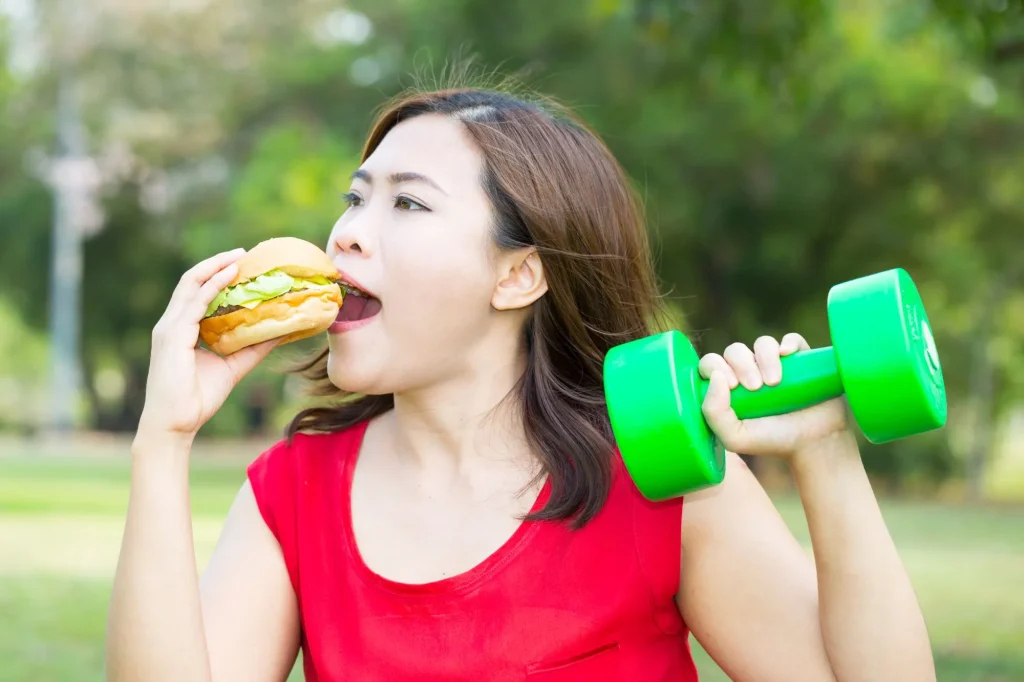 asain woman outside eating a burger with one hand and holding a hand weight in the other