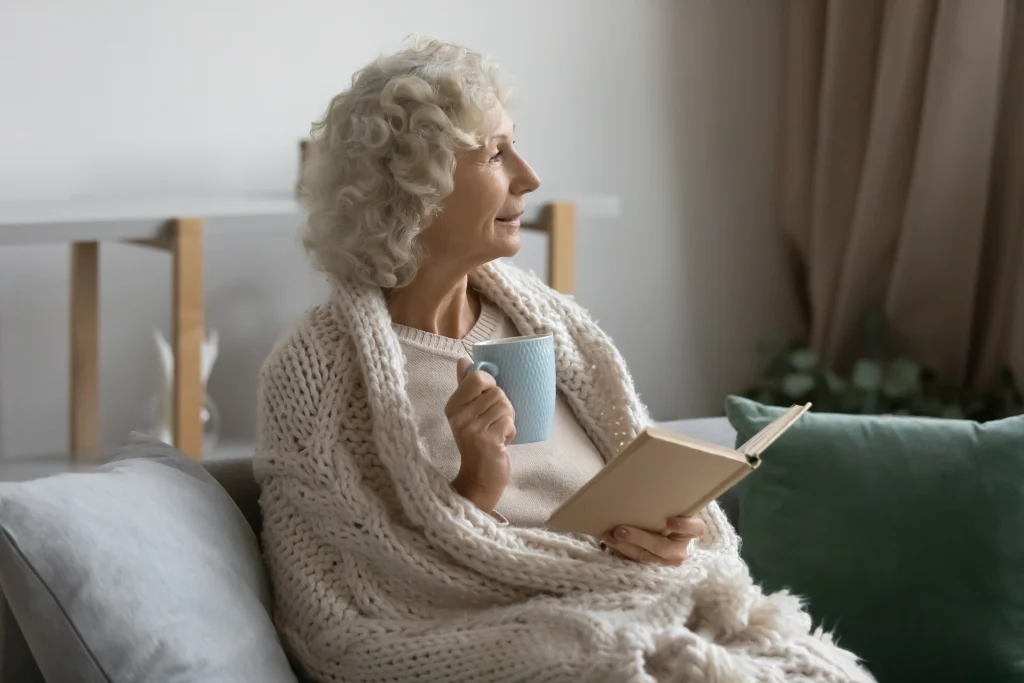 older senior woman reading a book and drinking coffee in bed