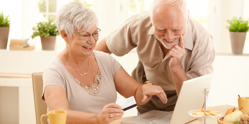 senior couple, man and a woman, sitting at a kitchen table looking at a computer screen together and smiling