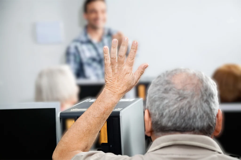 senior man raising hand in classroom setting with blurred people