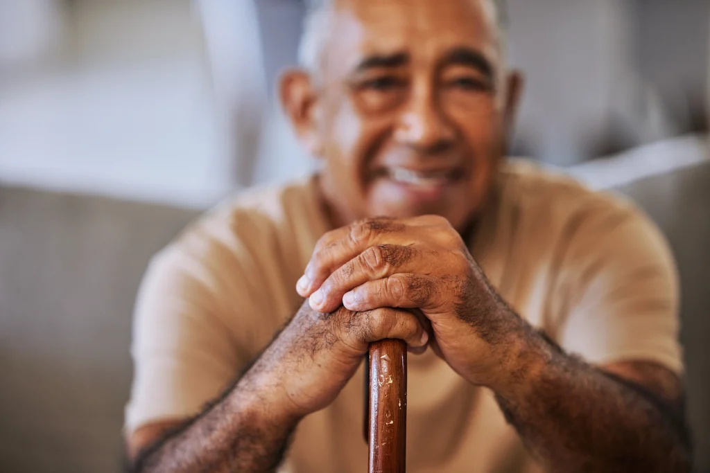 Portrait of a happy, kind black senior man hands with wrinkles, holding a walking stick and smiling in a retirement home sitting on outside relaxing