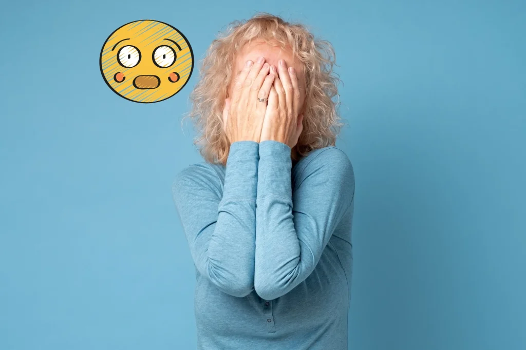embarrassed senior woman covering her face on a blue background with an embarassed emoji hovering over her