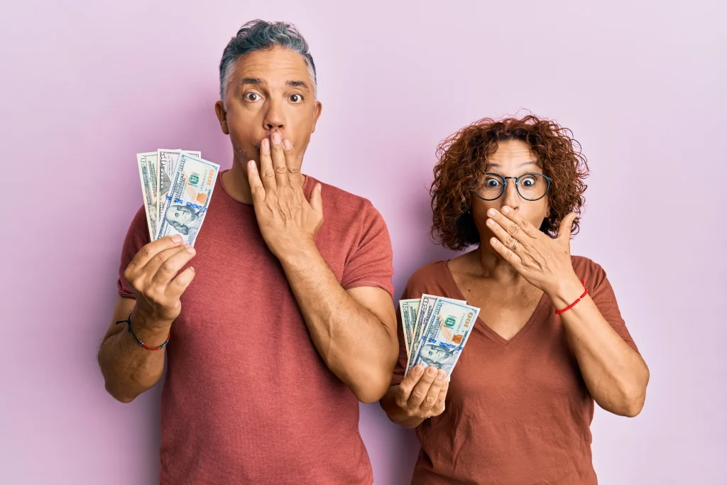 Beautiful middle age couple holding dollars covering mouth with hand, shocked and afraid for mistake. surprised expression
