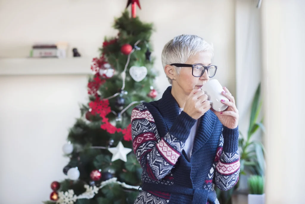 older senior woman standing in front of a christmas tree drinking from a mug