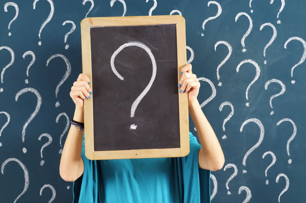 woman standing in front of a chalk board with lots of question marks and holding a large chalk board in front of her face with a large question mark