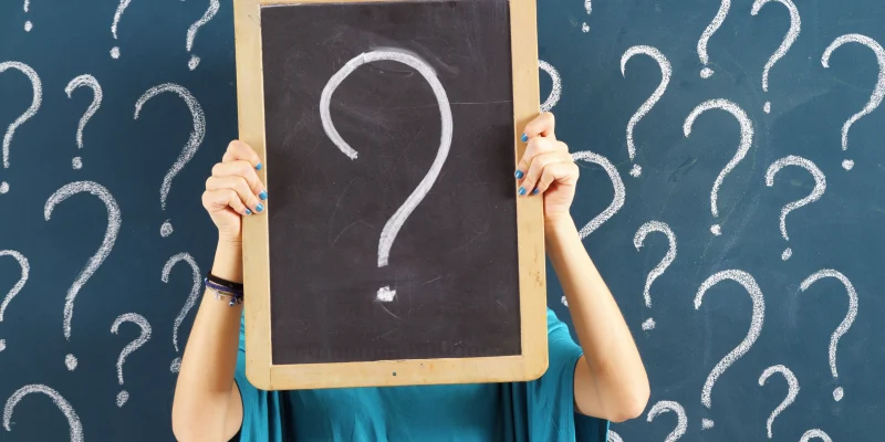woman standing in front of a chalk board with lots of question marks and holding a large chalk board in front of her face with a large question mark