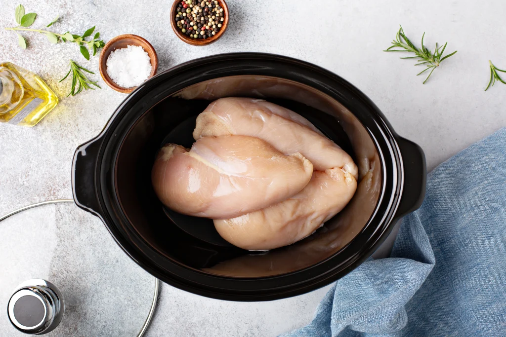 raw chicken in a slow cooker with ingredients on the counter nearby