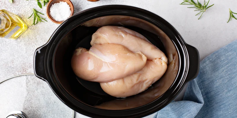 raw chicken in a slow cooker with ingredients on the counter nearby