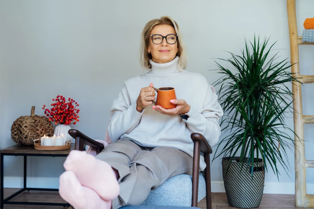 Middle aged woman relaxing with cup of hot drink in scandy style hygge interior home with fall mood interior decor. Lady dreaming, enjoy calm mood without stress, wellbeing alone. Cozy autumn at home.
