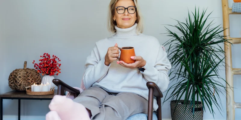 Middle aged woman relaxing with cup of hot drink in scandy style hygge interior home with fall mood interior decor. Lady dreaming, enjoy calm mood without stress, wellbeing alone. Cozy autumn at home.