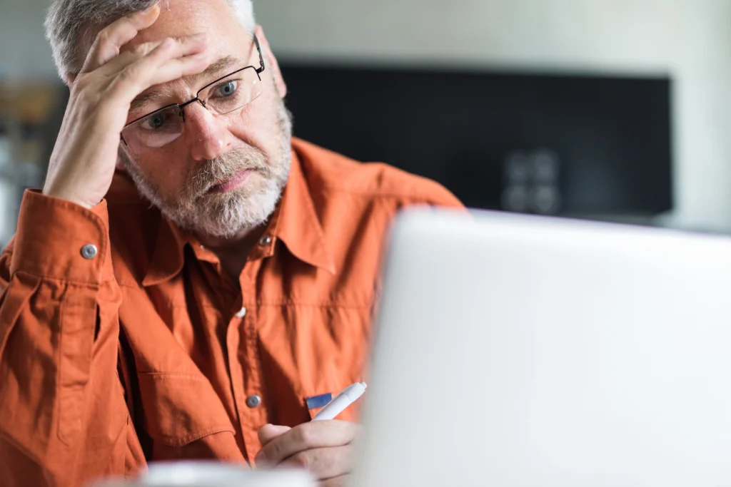 worried senior man wearing an orange button up shirt and using a computer looking at 2025 Medicare costs