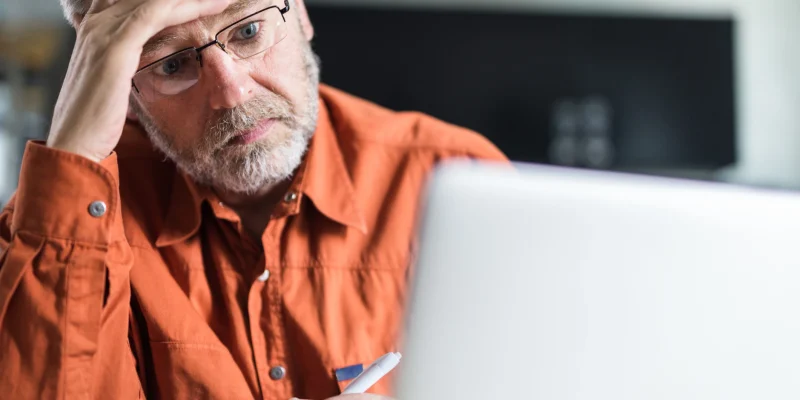 worried senior man wearing an orange button up shirt and using a computer